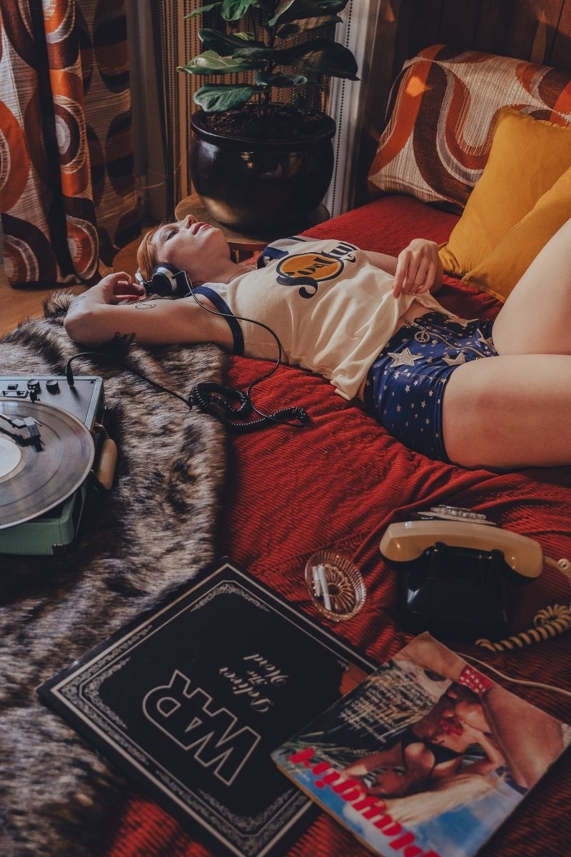Person lying on a bed wearing a soulful tank top with a red blanket, in a 1970s styled setting surrounded by books and a plant in a room with patterned curtains.