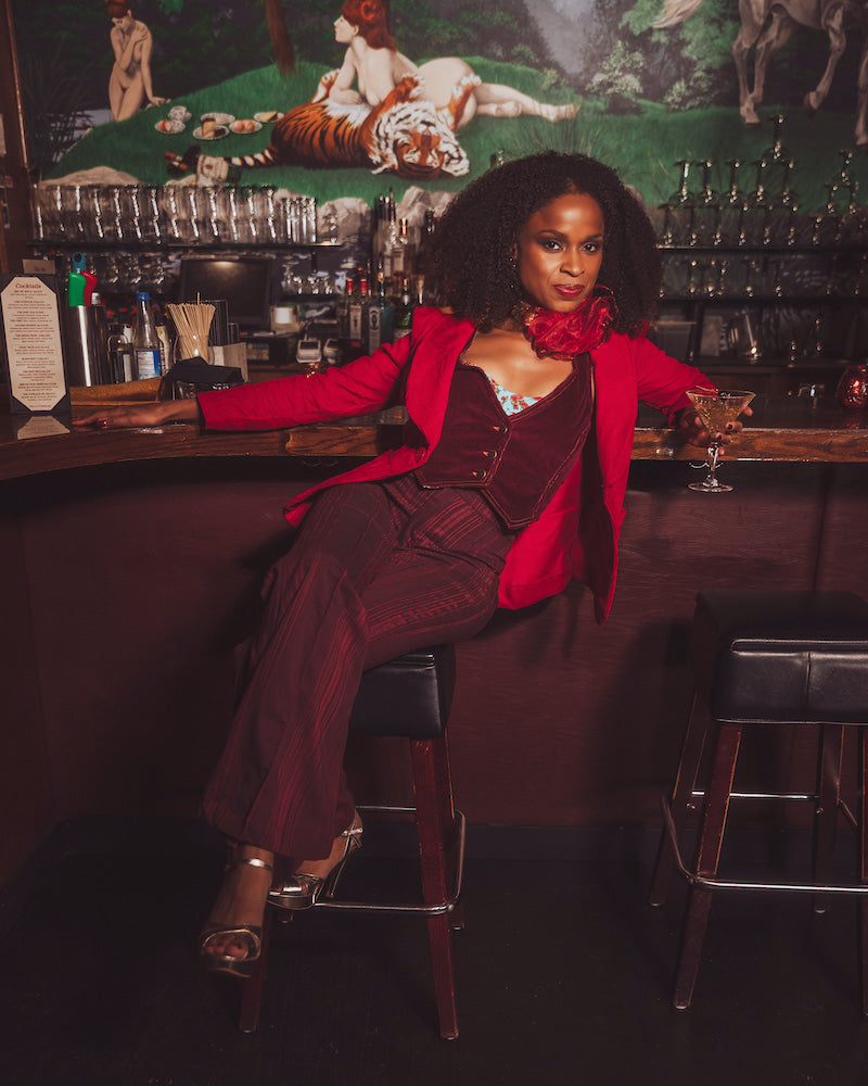 Woman in a red jacket and maroon suit sitting at a bar with a decorative wall in the background.