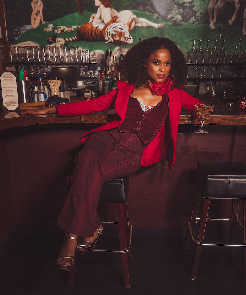 Woman in a red jacket and maroon suit sitting at a bar with a decorative wall in the background.