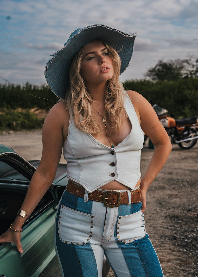 Woman in a white top and blue jeans standing next to a green car with a motorcycle in the background. Wearing Soul Sister since 1969
