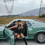 Woman in a black outfit sitting in a green car with power lines and mountains in the background wearing Soul Sister since 1969