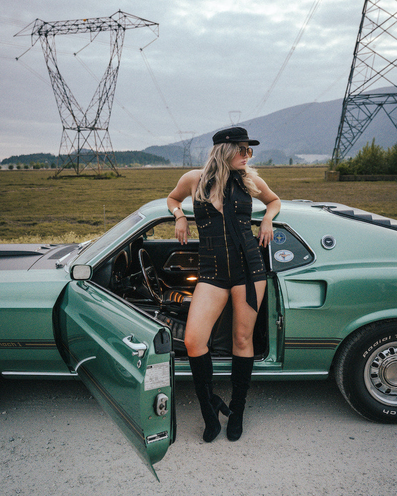 Woman in a black dress and boots standing next to a green car with power lines and mountains in the background wearing Soul Sister since 1969.
