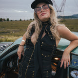 Woman in a black outfit with sunglasses leaning against a vintage car in an open field wearing Soul Sister since 1969.