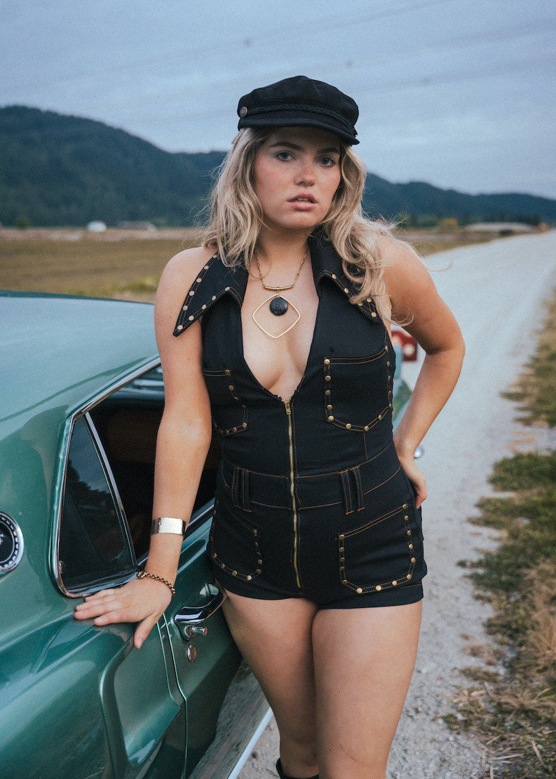Woman in a black outfit standing next to a vintage car on a rural road wearing Soul Sister since 1969.