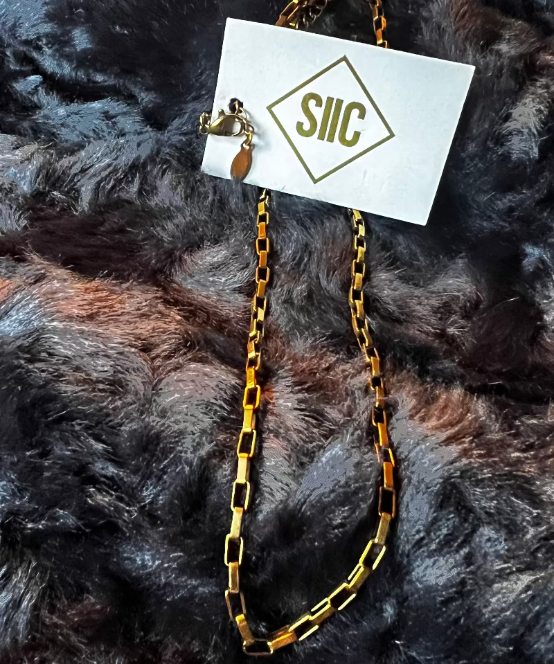 Gold chain necklace on a card with 'SILC' branding, placed on a textured dark fabric background.