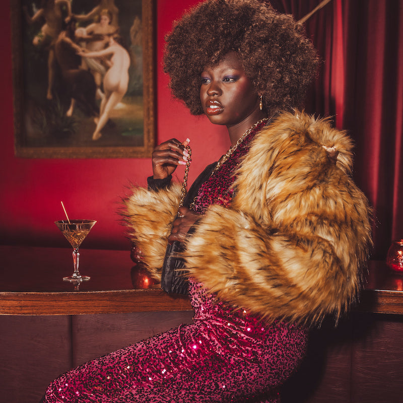 Woman in a glamorous setting with a 1970s style fur coat and cocktail, against a red wall with a painting.