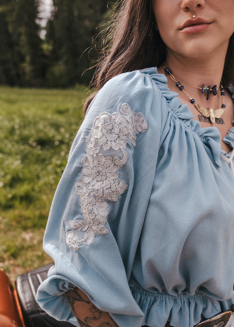 Close-up of a person wearing a light blue blouse with lace sleeves  designed by Soul Sister since 1969in an outdoor setting.