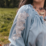Close-up of a person wearing a light blue blouse with lace sleeves  designed by Soul Sister since 1969in an outdoor setting.