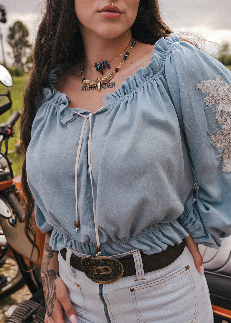 Person wearing a light blue blouse with ruffled sleeves  by Soul Sister since 1969, and a tattoo on the neck, standing next to a motorcycle.