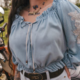Person wearing a light blue blouse with ruffled sleeves  by Soul Sister since 1969, and a tattoo on the neck, standing next to a motorcycle.