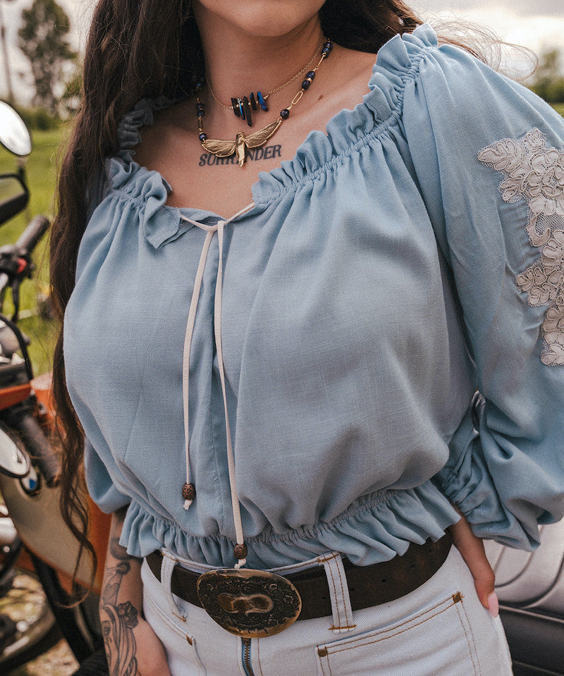 Person wearing a light blue blouse with ruffled sleeves  by Soul Sister since 1969, and a tattoo on the neck, standing next to a motorcycle.