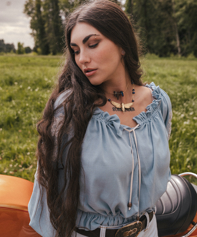 Woman in a light blue blouse by Soul Sister since 1969 sitting on a motorcycle in a grassy field with trees in the background
