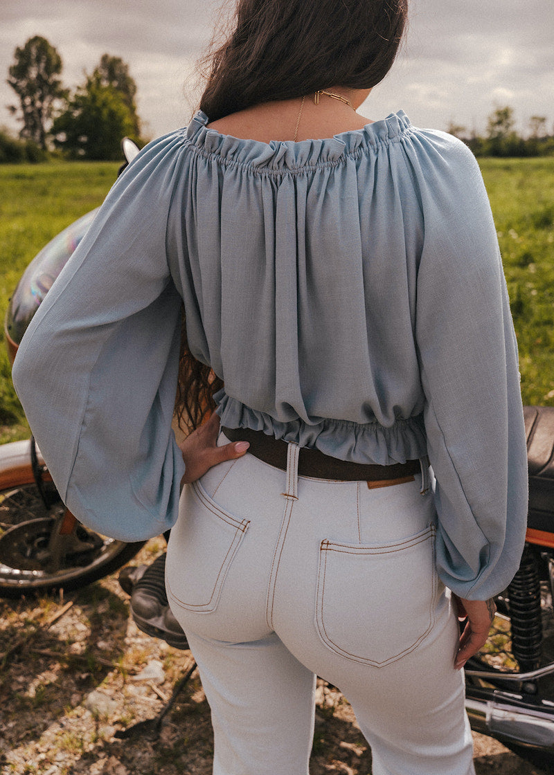 Person wearing a light blue blouse and sun faded denim bell bottom jeans by Soul Sister since 1969 standing next to a motorcycle in an open field.