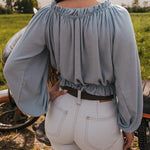 Person wearing a light blue blouse and sun faded denim bell bottom jeans by Soul Sister since 1969 standing next to a motorcycle in an open field.