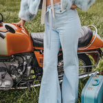 Person wearing a light blue fur coat and jeans standing next to an orange motorcycle with a teal suitcase on grass wearing Soul Sister since 1969.