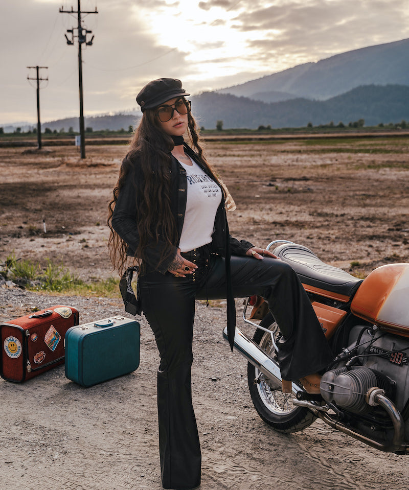 Woman sitting on a motorcycle with suitcases nearby, set against a scenic background wearing Soul Sister since 1969.