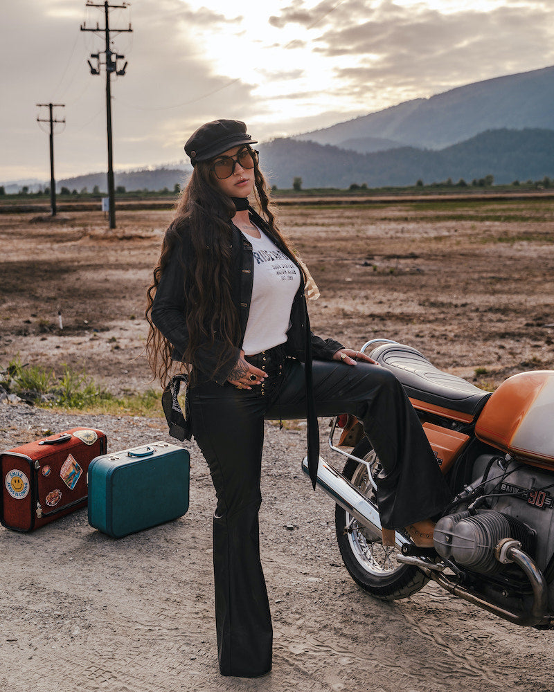 Woman sitting on a motorcycle with suitcases nearby, set against a scenic background wearing clothing designed and made by Soul Sister since 1969..