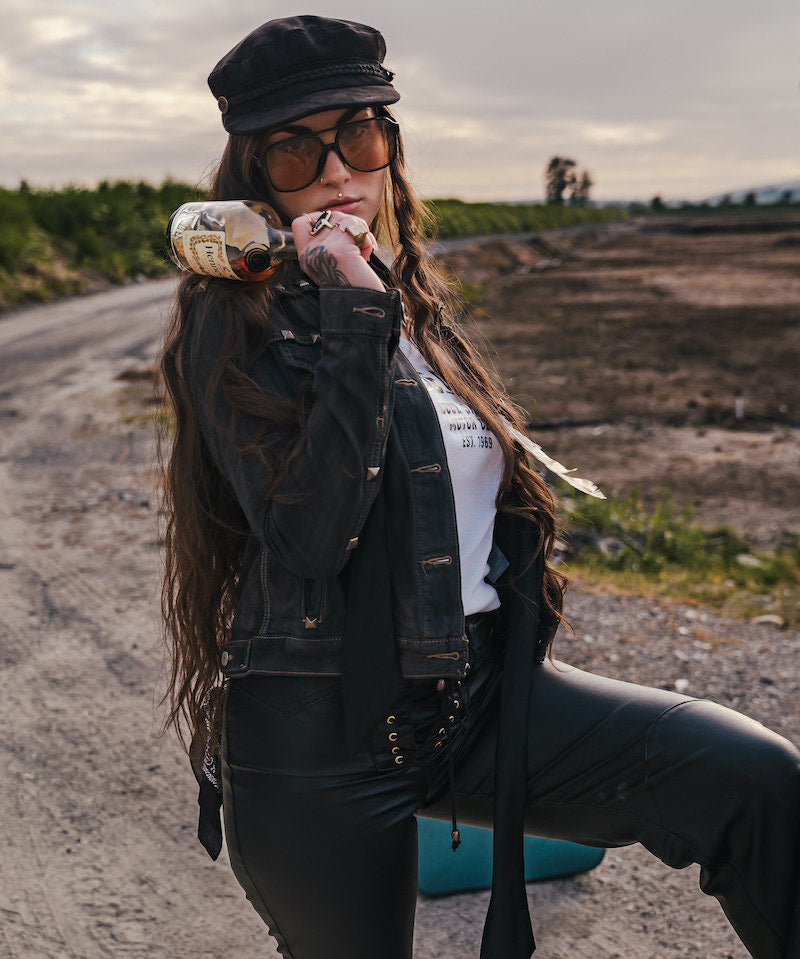 Woman in a black jacket and hat holding a bottle in an open field wearing Soul Sister since 1969.