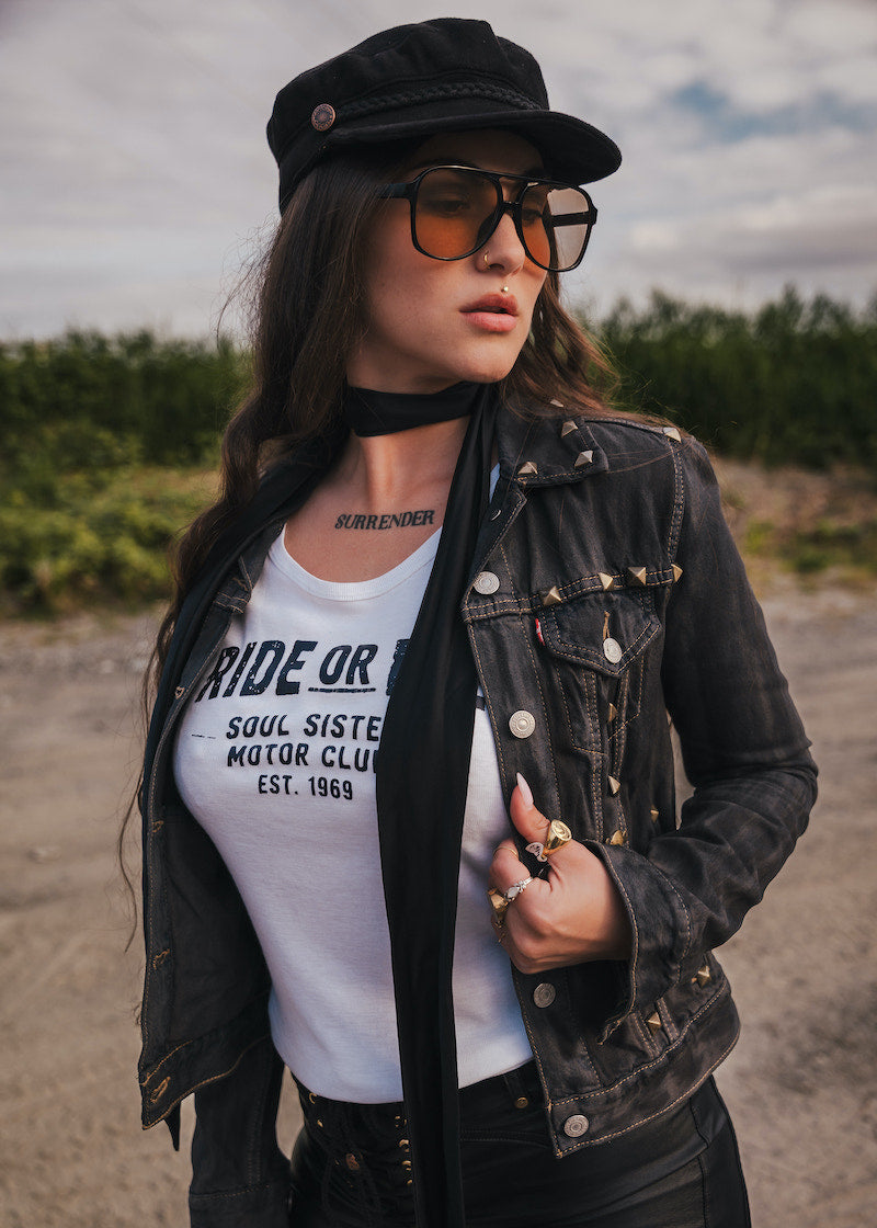 Woman wearing a black jacket, white t-shirt, and sunglasses  and a black hat outdoors wearing Soul Sister since 1969.