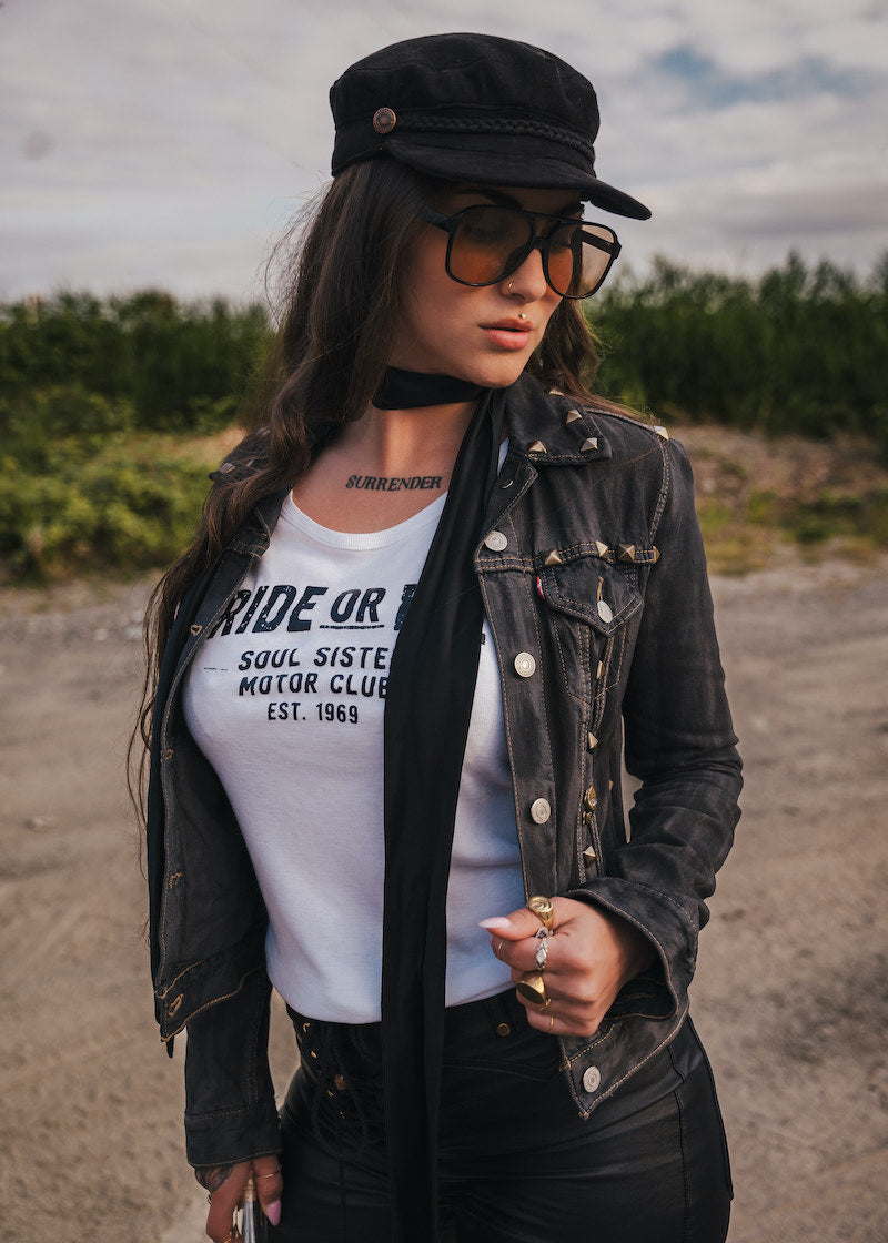 Woman wearing a black jacket, white t-shirt with text, and sunglasses outdoors wearing Soul Sister since 1969.