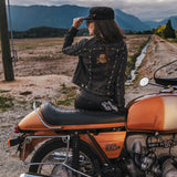 Person sitting on an orange motorcycle in a scenic outdoor setting with mountains in the background wearing Soul Sister since 1969