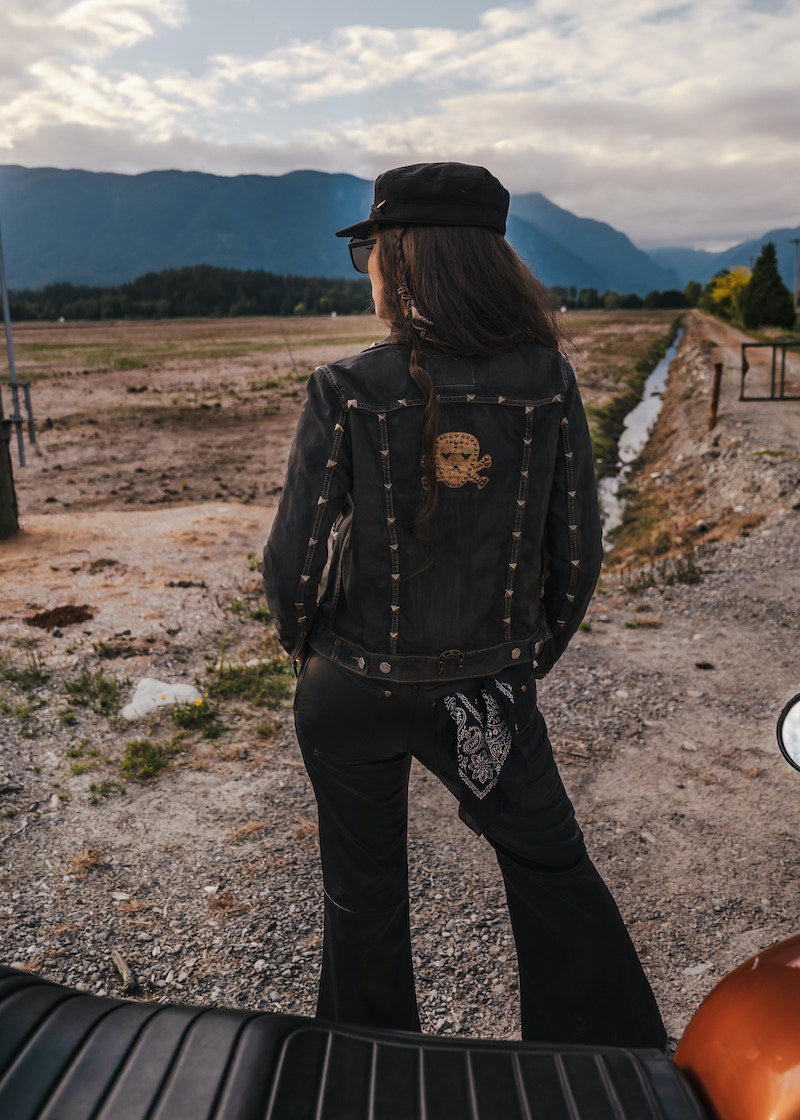 Person wearing a black jacket with skull design and cap, standing on a dirt road with mountains in the background wearing Soul Sister since 1969.