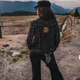 Person wearing a black jacket with skull design and cap, standing on a dirt road with mountains in the background wearing Soul Sister since 1969.