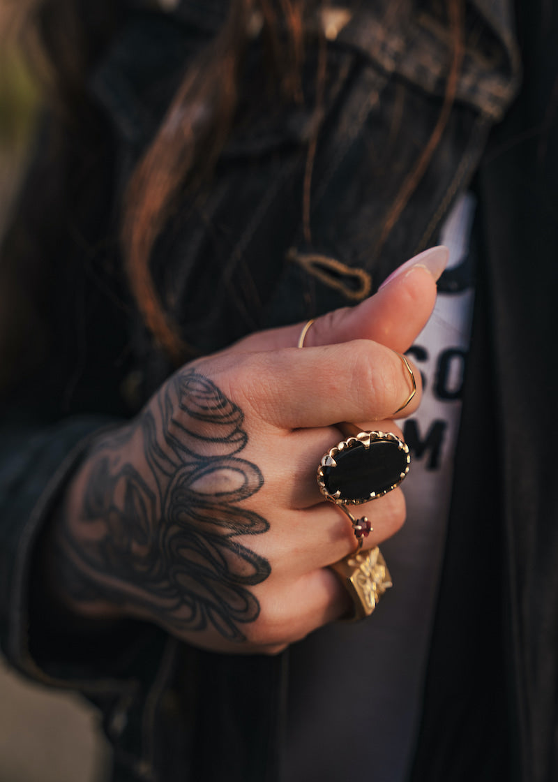 Hand with tattoos and multiple rings, wearing a dark jacket wearing clothing designed and made by Soul Sister since 1969..