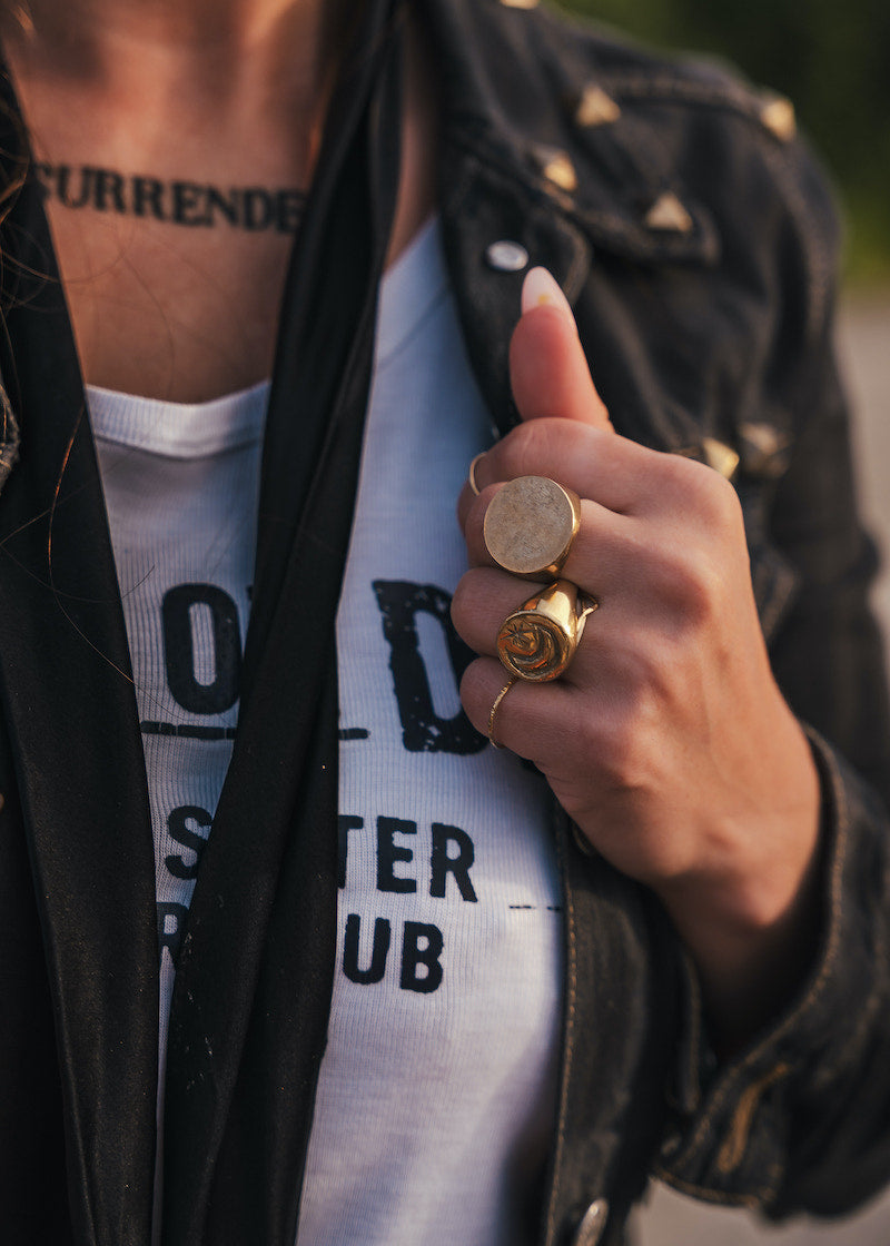 Person wearing a black jacket with gold buttons, white shirt with text, and multiple rings on a blurred background wearing Soul Sister since 1969.