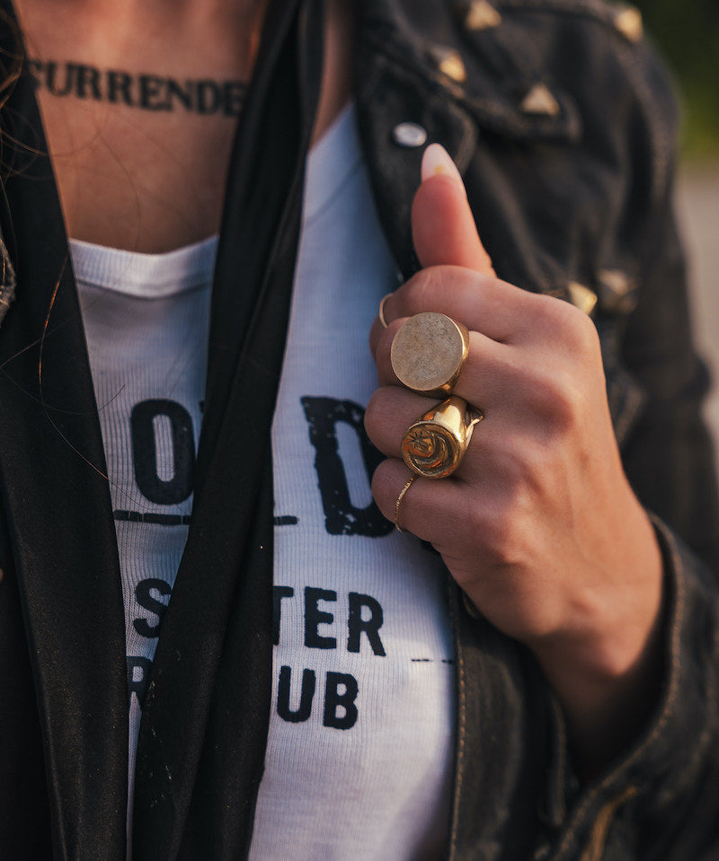 Person wearing a black jacket with gold buttons, white shirt with text, and multiple rings on a blurred background wearing clothing designed and made by Soul Sister since 1969..