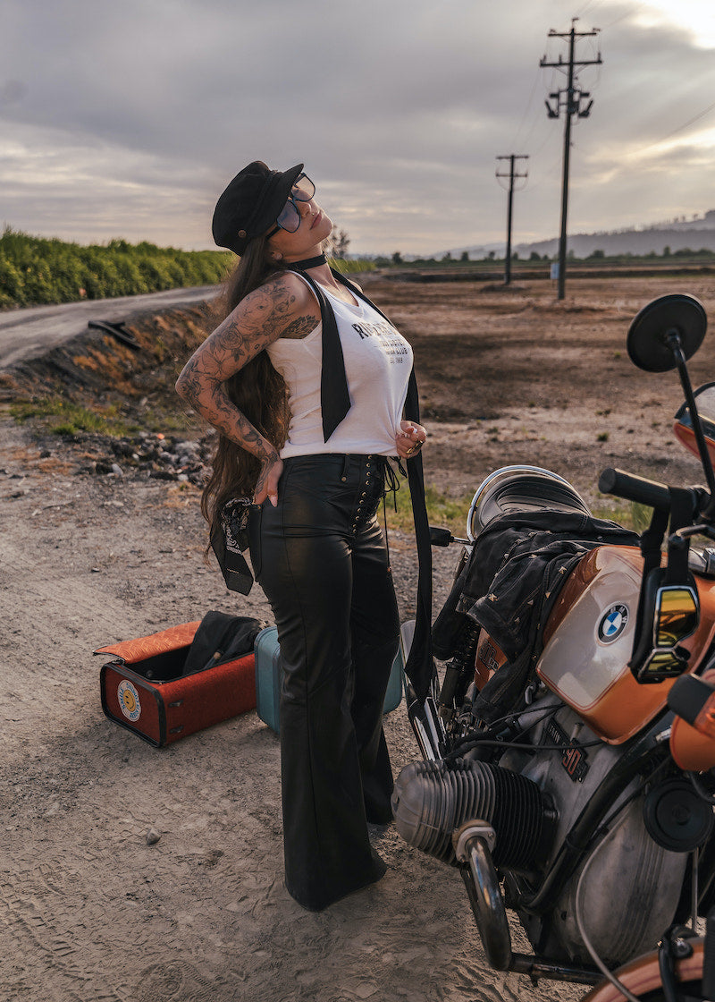 Woman in a white tank top and black pants standing next to a motorcycle on a dirt road wearing clothing designed and made by Soul Sister since 1969.