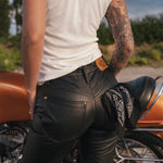 Person wearing black leather pants and a white tank top standing next to a motorcycle wearing clothing designed and made by Soul Sister since 1969..