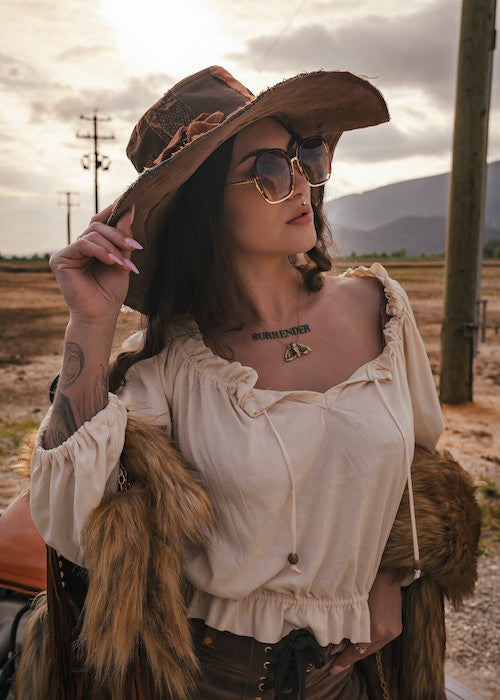 Woman in an oversized hat and sunglasses with a tattoo on her neck, standing in a field wearing Soul Sister since 1969.