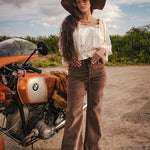 Woman in vintage attire standing next to a BMW motorcycle on a road with a scenic background wearing Soul Sister since 1969