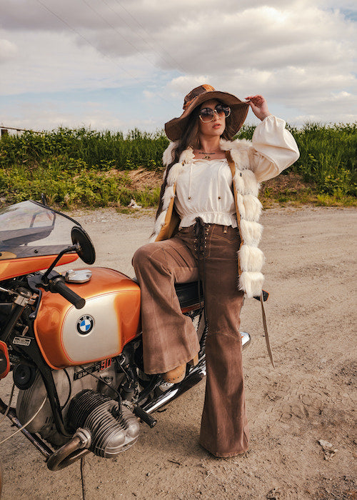 Person sitting on a BMW motorcycle wearing a white blouse, brown pants, and a hat with a fur-lined coat wearing Soul Sister since 1969.