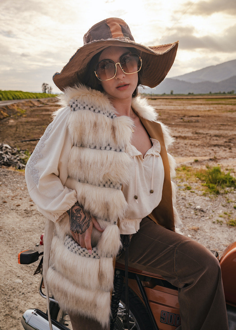 Person wearing a fur vest and wide-brimmed hat sitting on a motorcycle in an open field. Wearing Soul Sister since 1969