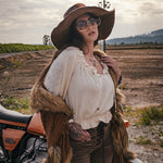 Person in rustic attire with a fur vest and wide-brimmed hat standing next to a motorcycle in an open field. wearing Soul Sister since 1969