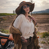 Person in rustic attire with a fur vest and wide-brimmed hat standing next to a motorcycle in an open field. wearing Soul Sister since 1969