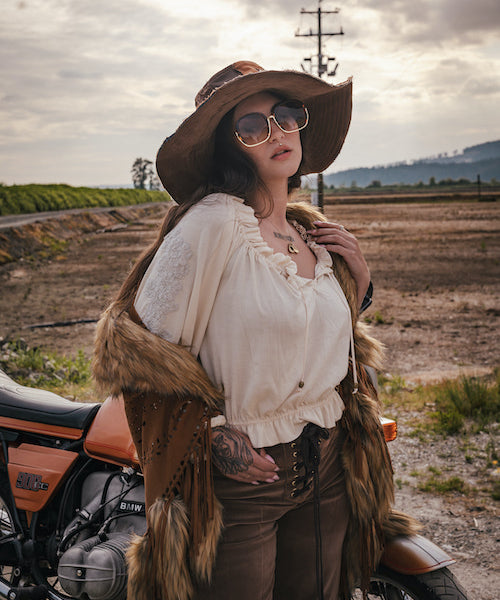 Person in rustic attire with a fur vest and wide-brimmed hat standing next to a motorcycle in an open field. wearing Soul Sister since 1969