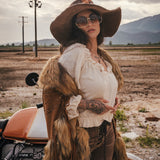 Woman in rustic attire with fur vest and wide-brimmed hat standing next to a motorcycle in an open field wearing Soul Sister since 1969.