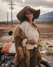 Woman in rustic attire with fur vest and wide-brimmed hat standing next to a motorcycle in an open field wearing Soul Sister since 1969.