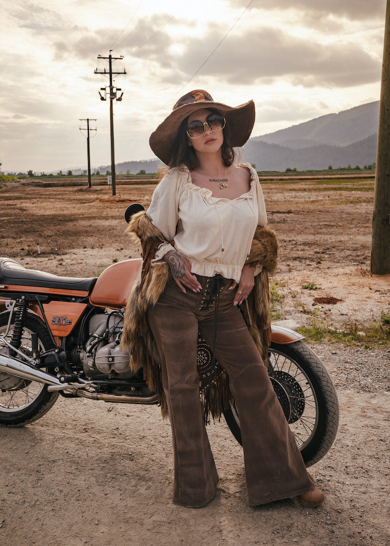 Person in vintage-style clothing standing next to a motorcycle in an open field with mountains in the background. wearing Soul Sister since 1969