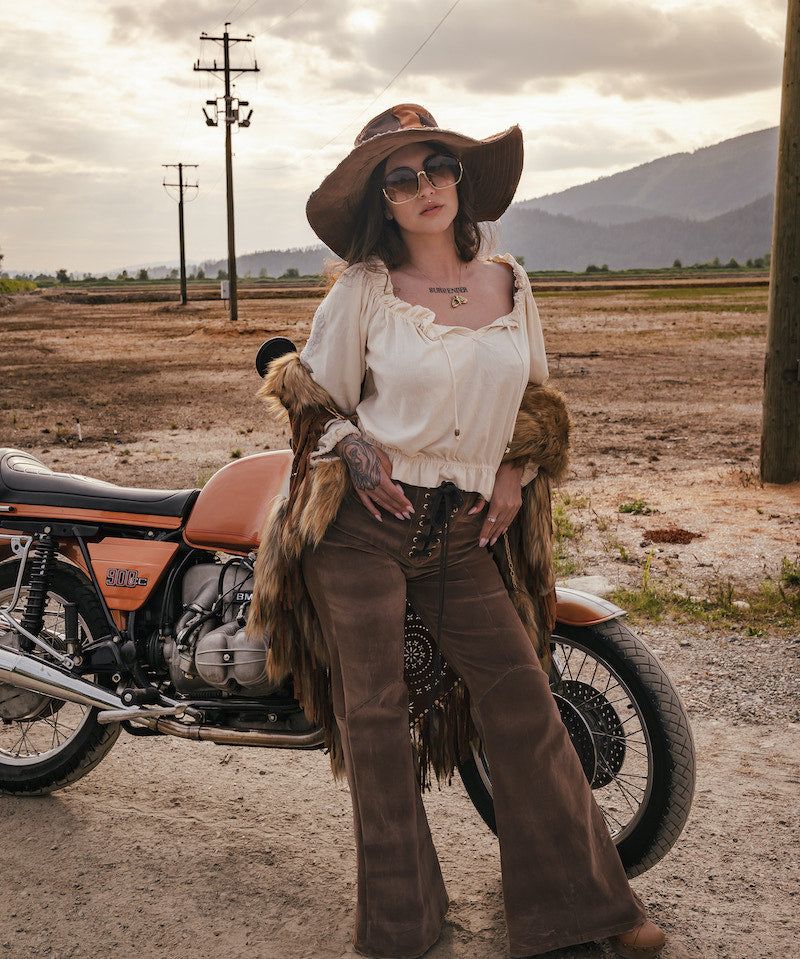 Person in vintage-style clothing standing next to a motorcycle in an open field with mountains in the background. wearing Soul Sister since 1969