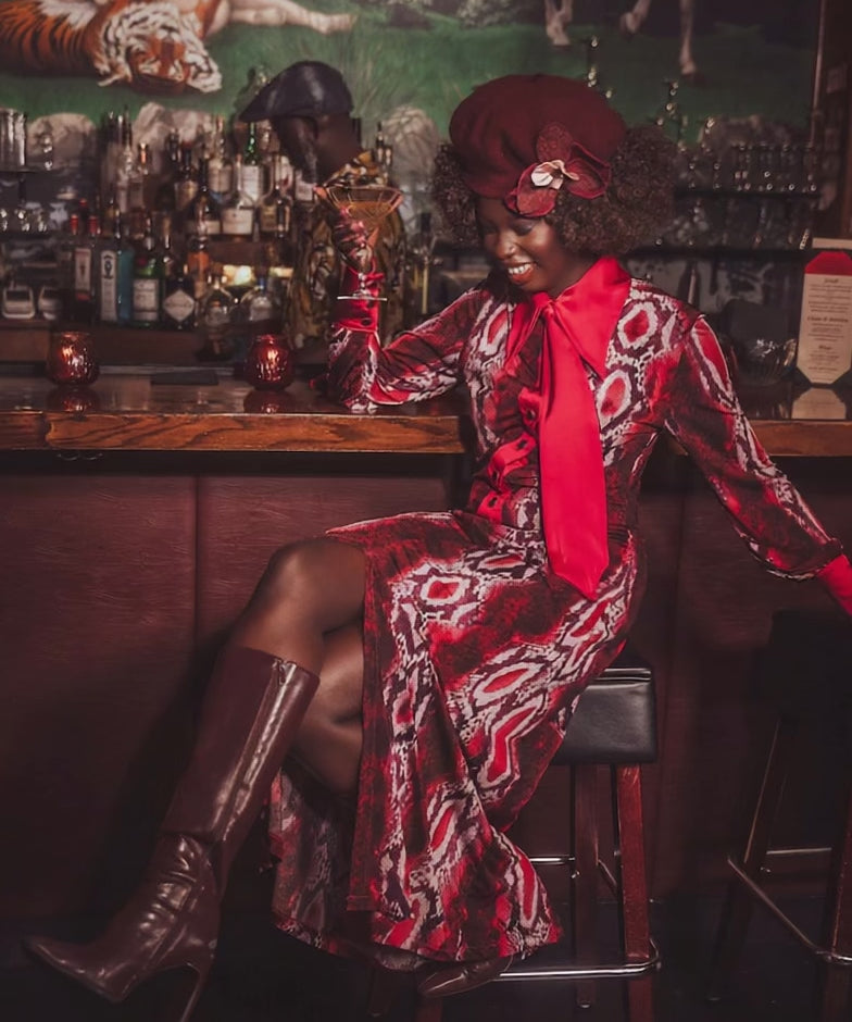 A woman sits at the bar with a martini in her hand in a speakeasy setting wearing a cool 70s inspired retro dress in snake print