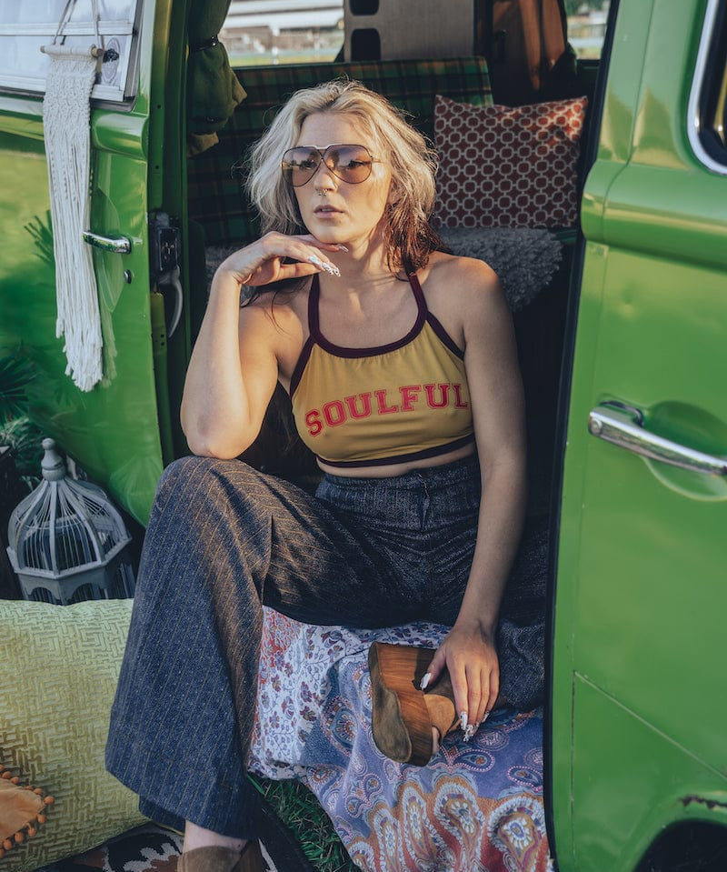 A person wearing a yellow halter top with the word 'SOUULFUL' printed on it, sitting in a green vehicle with their legs dangling outside.