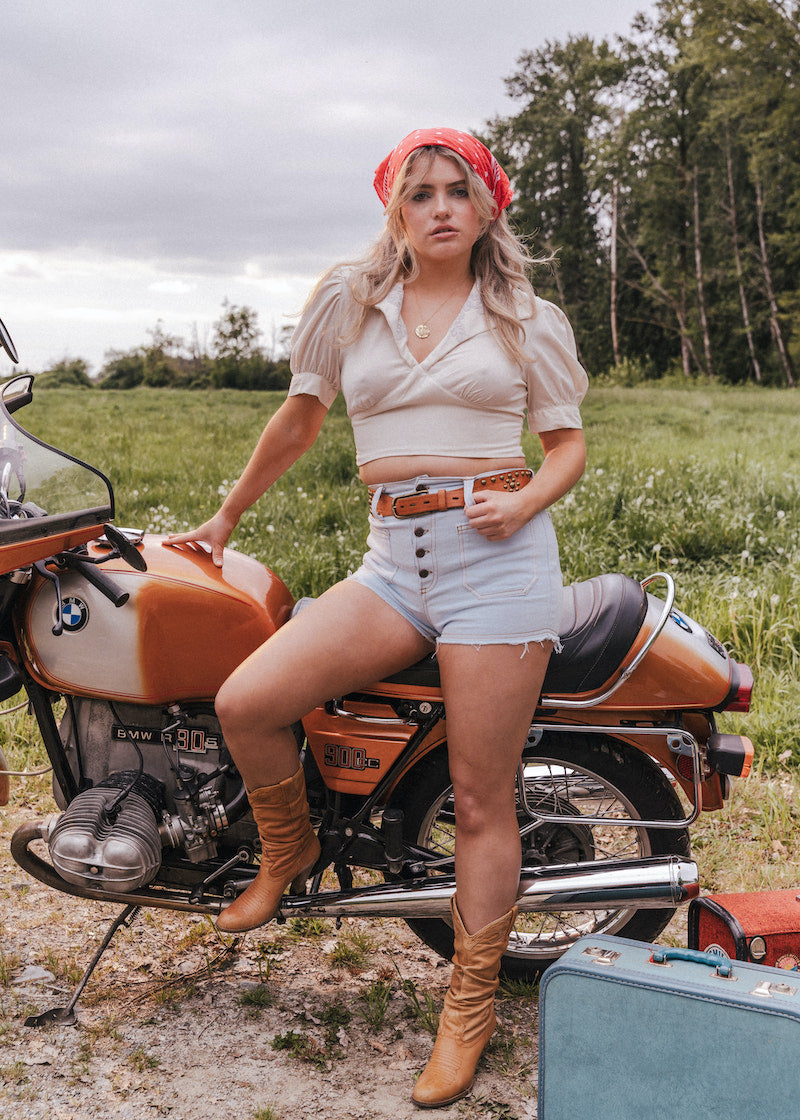 Woman sitting on a motorcycle in a field with a suitcase nearby wearing Soul Sister since 1969