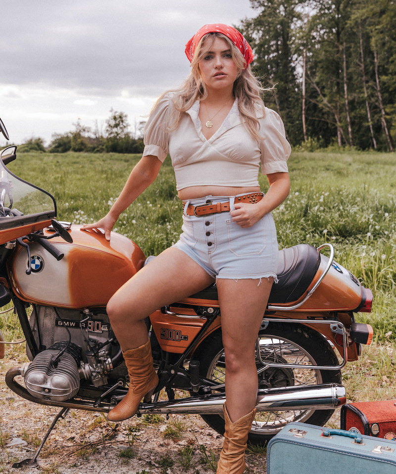 Woman sitting on a motorcycle in a field with a suitcase nearby wearing Soul Sister since 1969