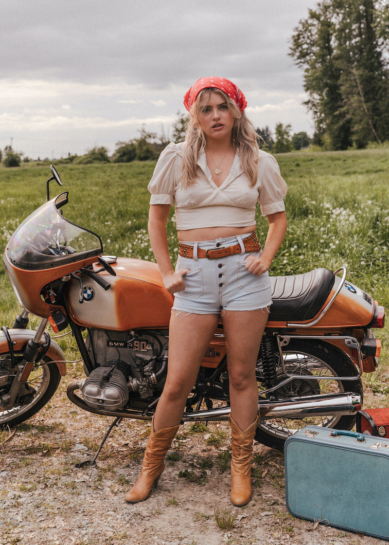Woman standing next to a vintage motorcycle in a field wearing Soul Sister since 1969.