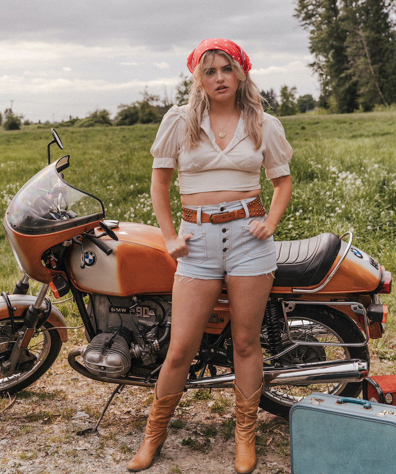 Woman standing next to a vintage motorcycle in a field wearing Soul Sister since 1969.